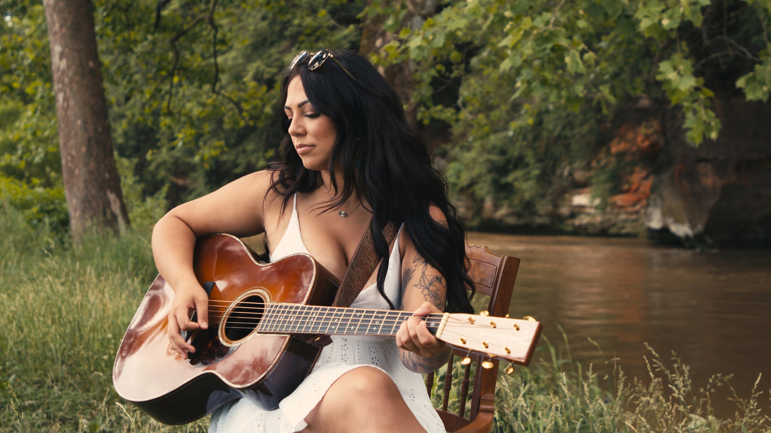 Booking Promo #8. Emily Raff playing an acoustic guitar by a river, surrounded by greenery.