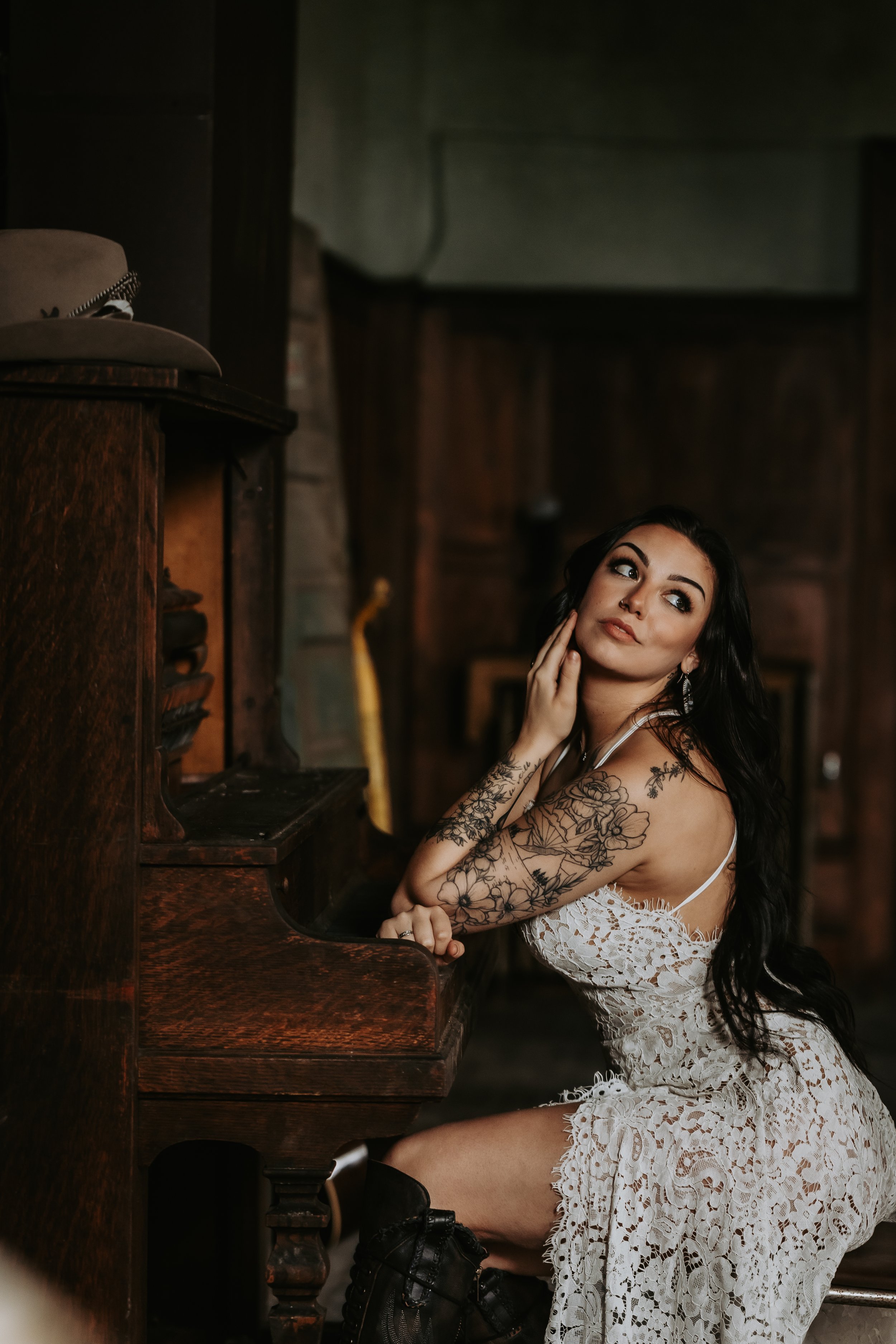 Booking Promo #5. Emily Raff with tattoos wearing a lace dress, sitting at an antique piano, posing thoughtfully.
