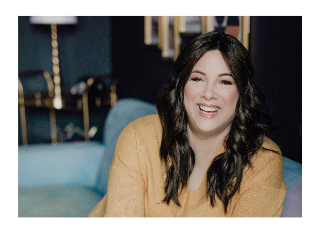 Smiling virtual assistant with dark hair sitting on a blue couch in a warmly lit room, ready to support you and your business.