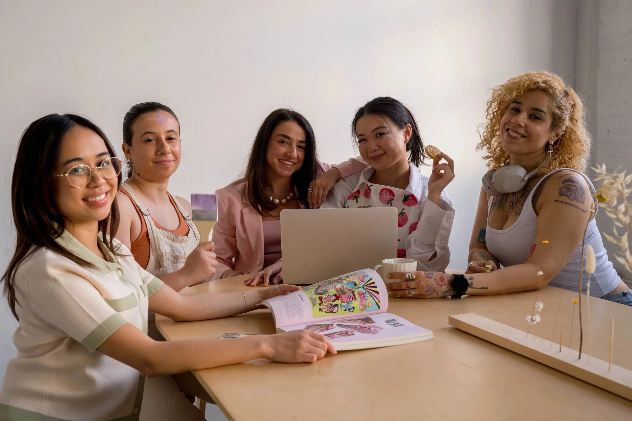 A diverse group of six women sitting at a wooden table in a bright room. They are smiling and looking at the camera, with some holding a magazine and a cookie. There is a laptop and a book on the table, and the background is plain with some subtle decoration.