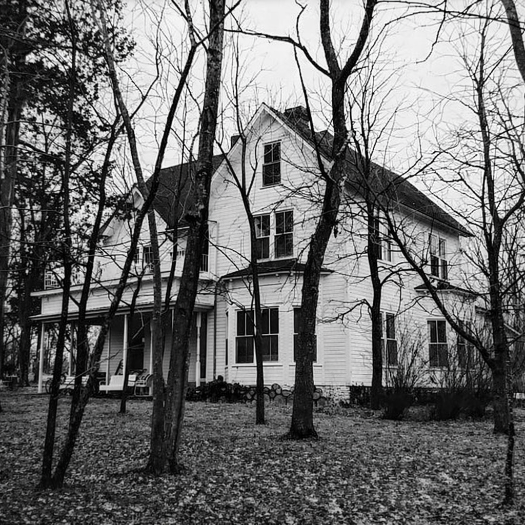 A large white house with multiple stories, surrounded by leafless trees, with a front porch and several windows, seen in a black and white photograph.