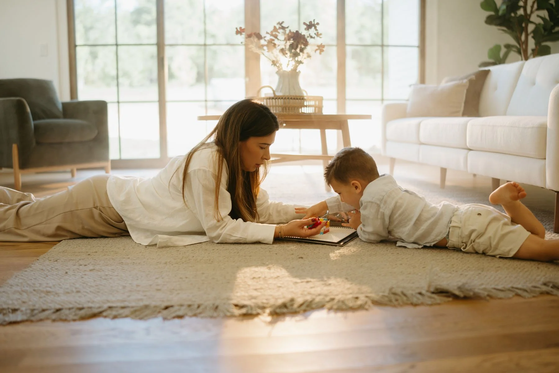 A woman and a young boy lying on a beige carpet in a bright living room, playing with toys and a tablet together.