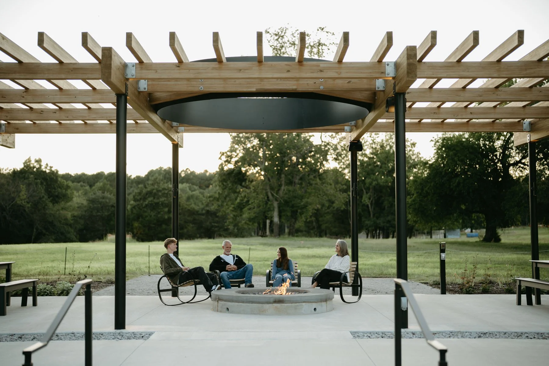 Group of four people sitting around a fire pit beneath a wooden pergola in a park with trees in the background