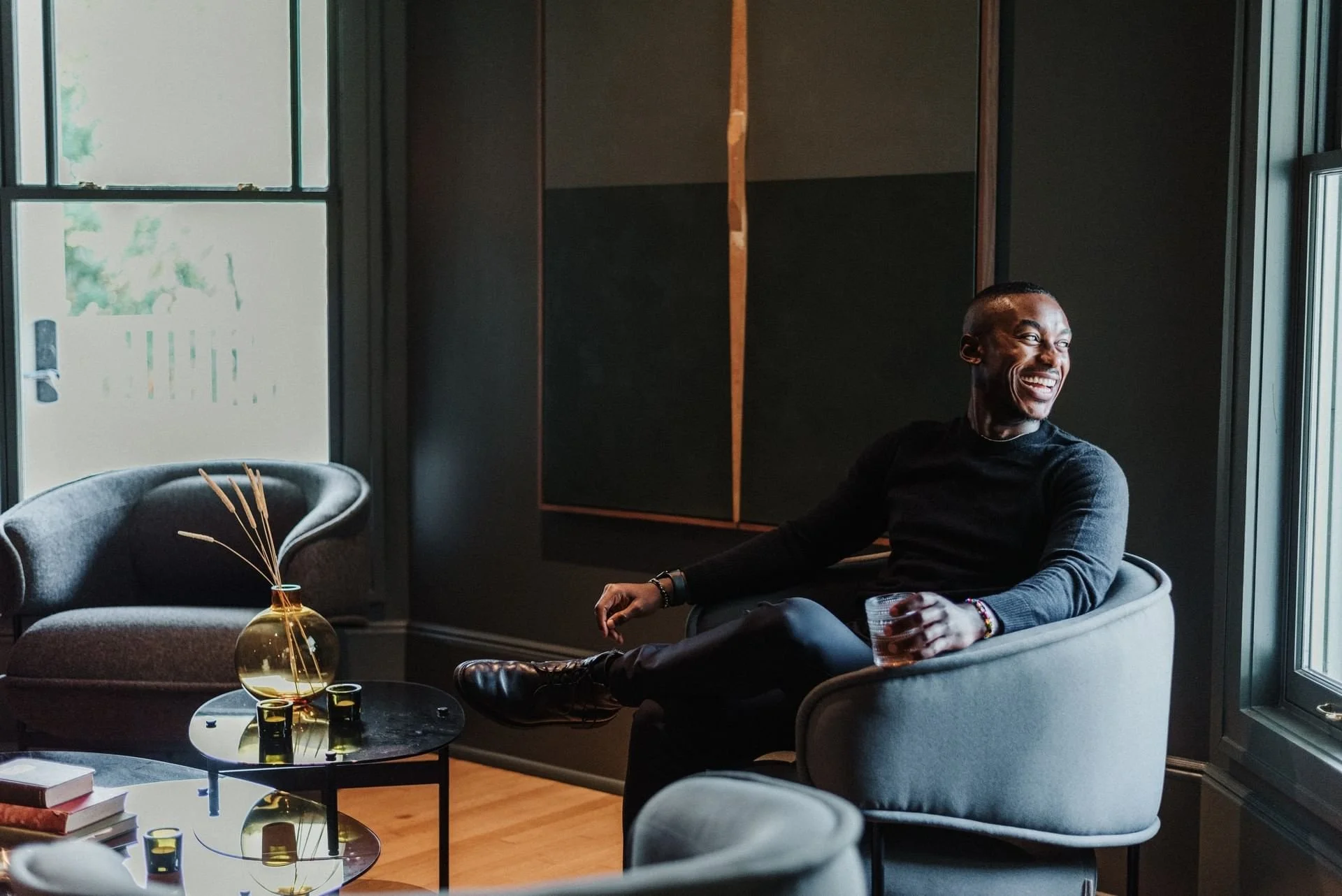 A man sitting on a stylish armchair in a modern living room, smiling and laughing, holding a glass in one hand, with a coffee table with a glass vase and books in the foreground, and large windows letting in natural light.