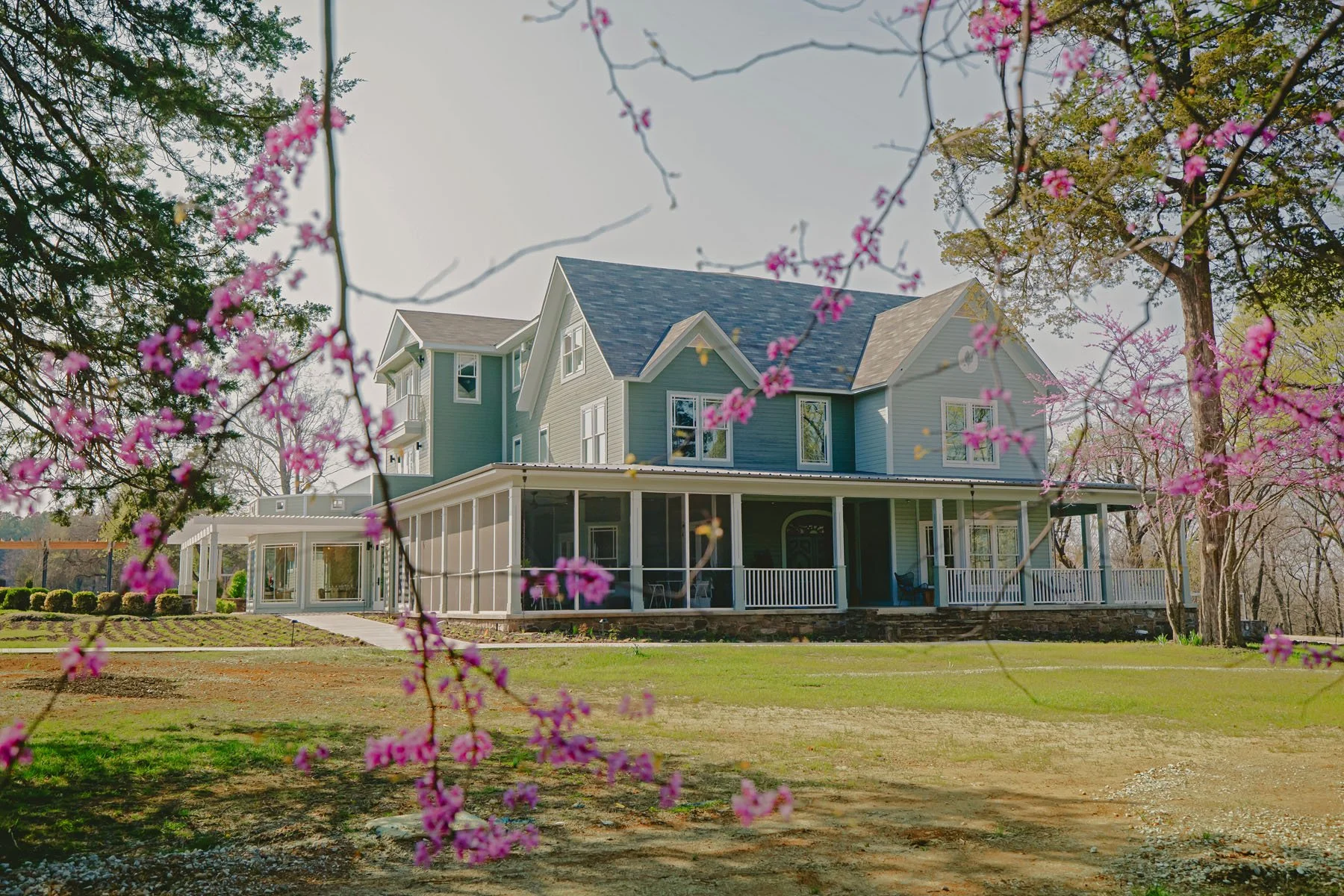 A large two-story house with a wraparound porch, painted in shades of blue and white, surrounded by pink flowering trees and green grass, with a partly cloudy sky in the background.