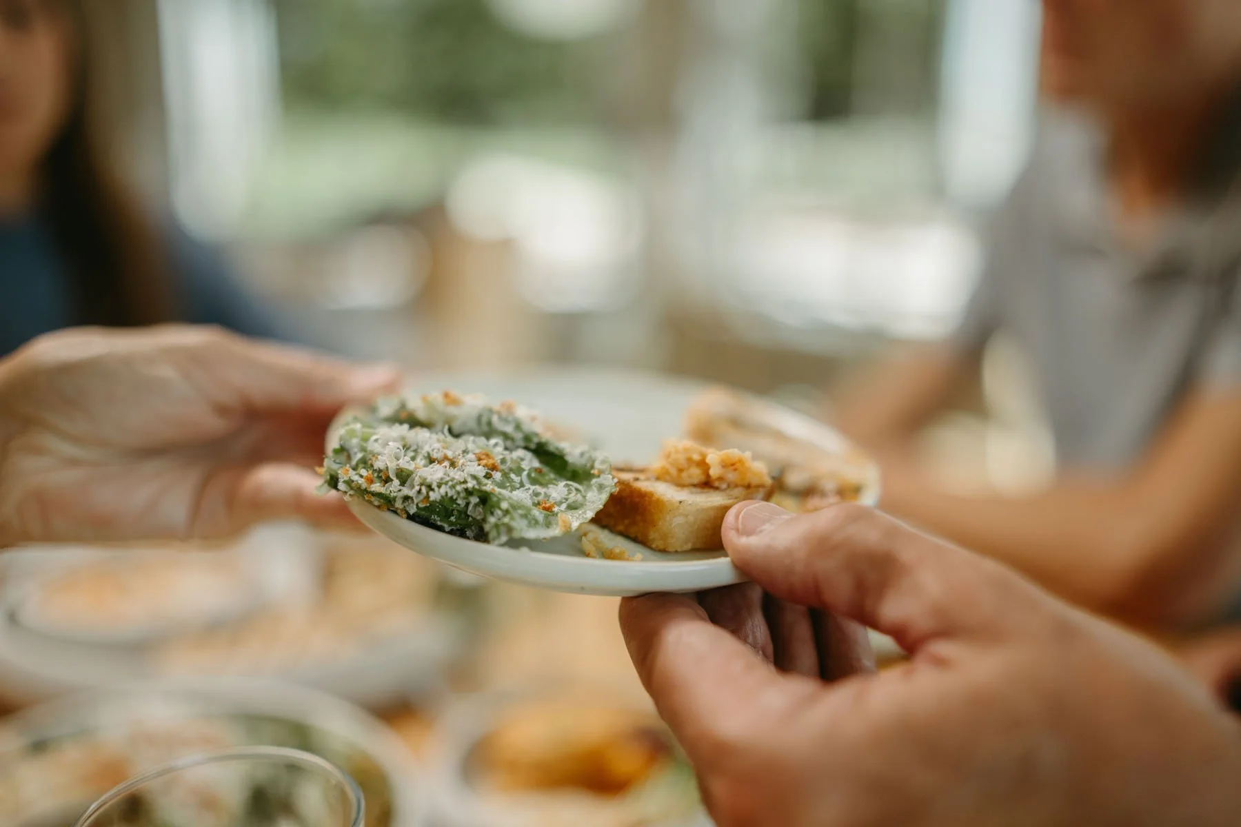 Hands holding a small plate with two pieces of bread topped with spreads or toppings, blurred background of more food dishes and a person in a casual setting.