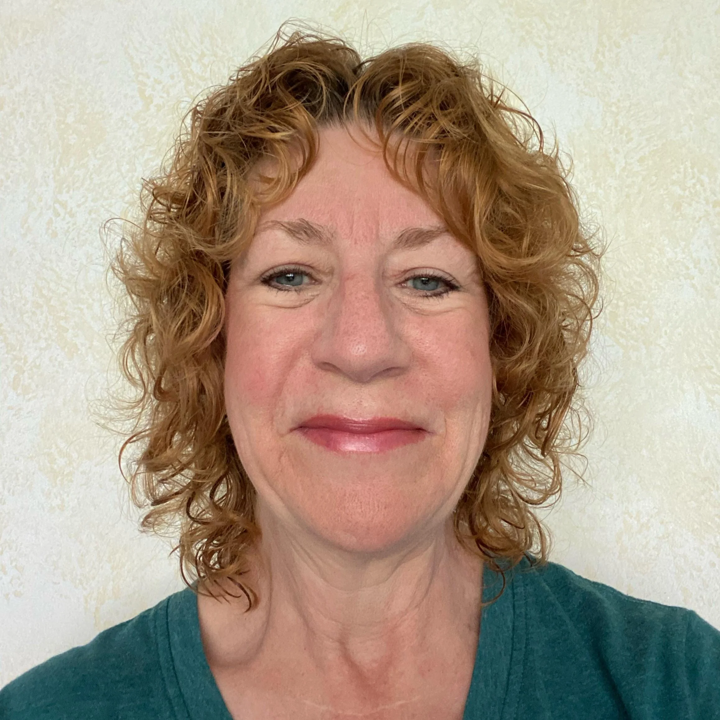Close-up of a woman with curly red hair, light blue eyes, and a slight smile, wearing a teal shirt, against a textured cream-colored wall.