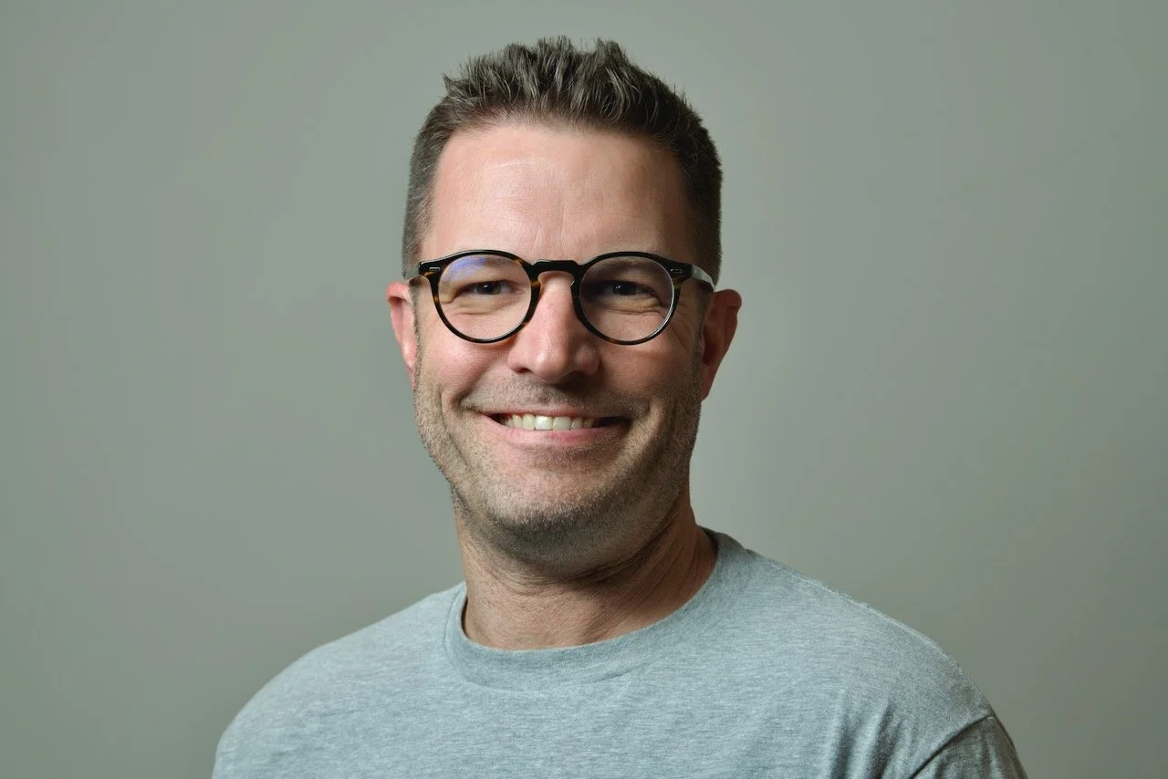 A smiling man with glasses and short brown hair, wearing a light grey shirt, standing against a plain grey background.