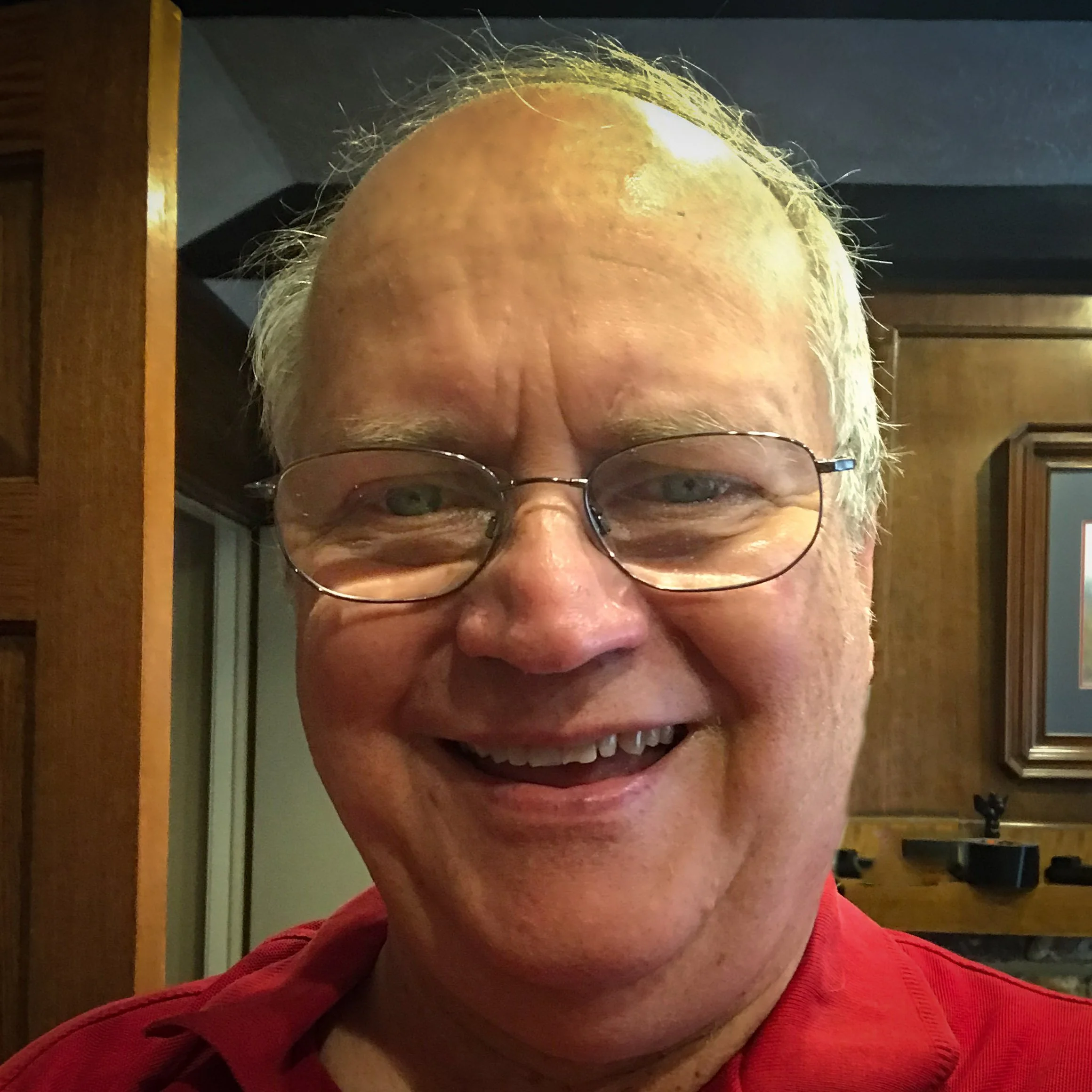 A smiling middle-aged man with glasses wearing a red shirt in a room with wooden paneling and framed artwork.