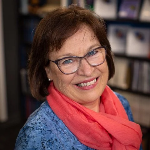 A smiling woman with short brown hair, glasses, wearing a blue patterned top and a pink scarf, standing in front of bookshelves.