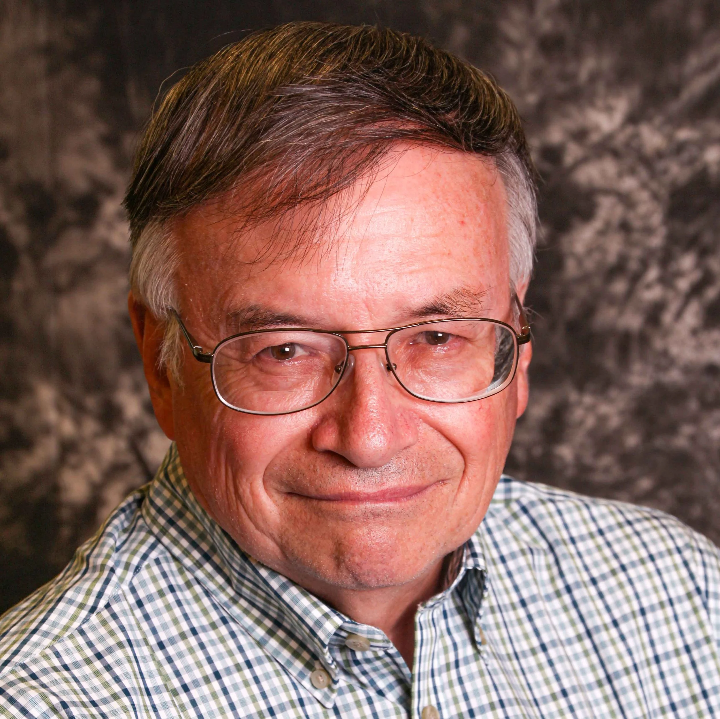 Close-up portrait of an older man with glasses, smiling, wearing a checkered shirt, with a blurred dark background.