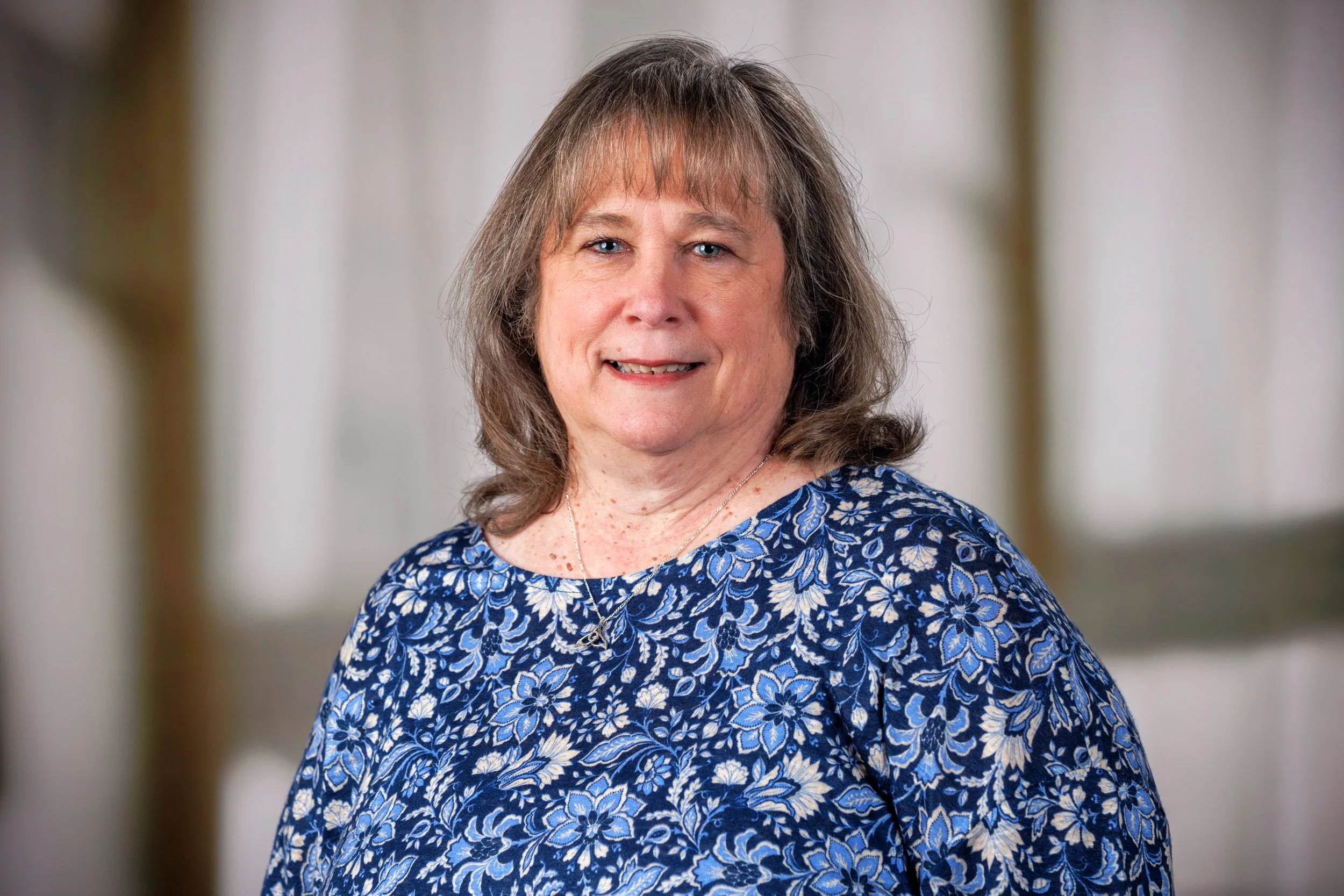 Portrait of an older woman with shoulder-length gray hair, wearing a blue floral blouse, smiling, with a neutral background.