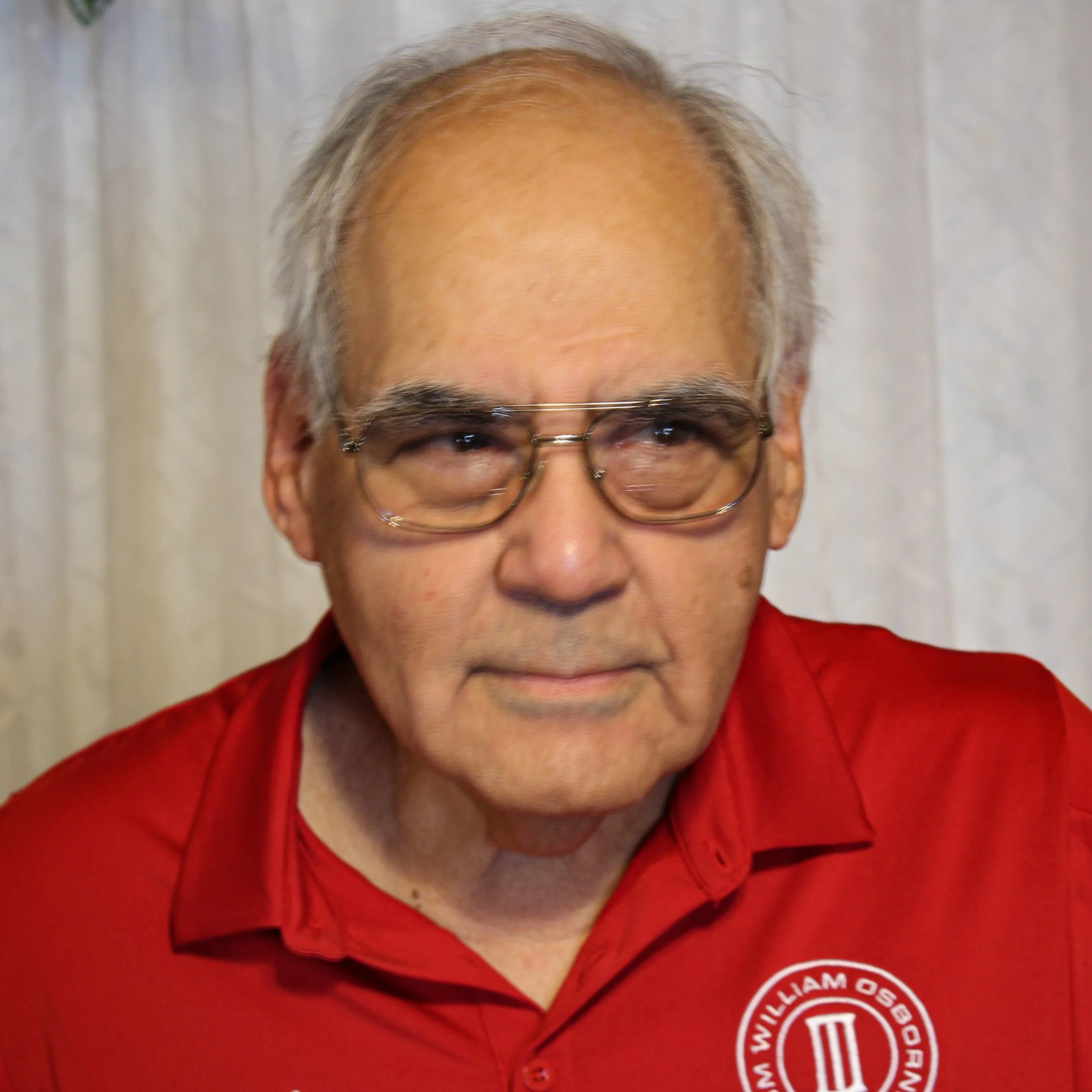 An elderly man with glasses wearing a red shirt with a William O'Sullivan logo