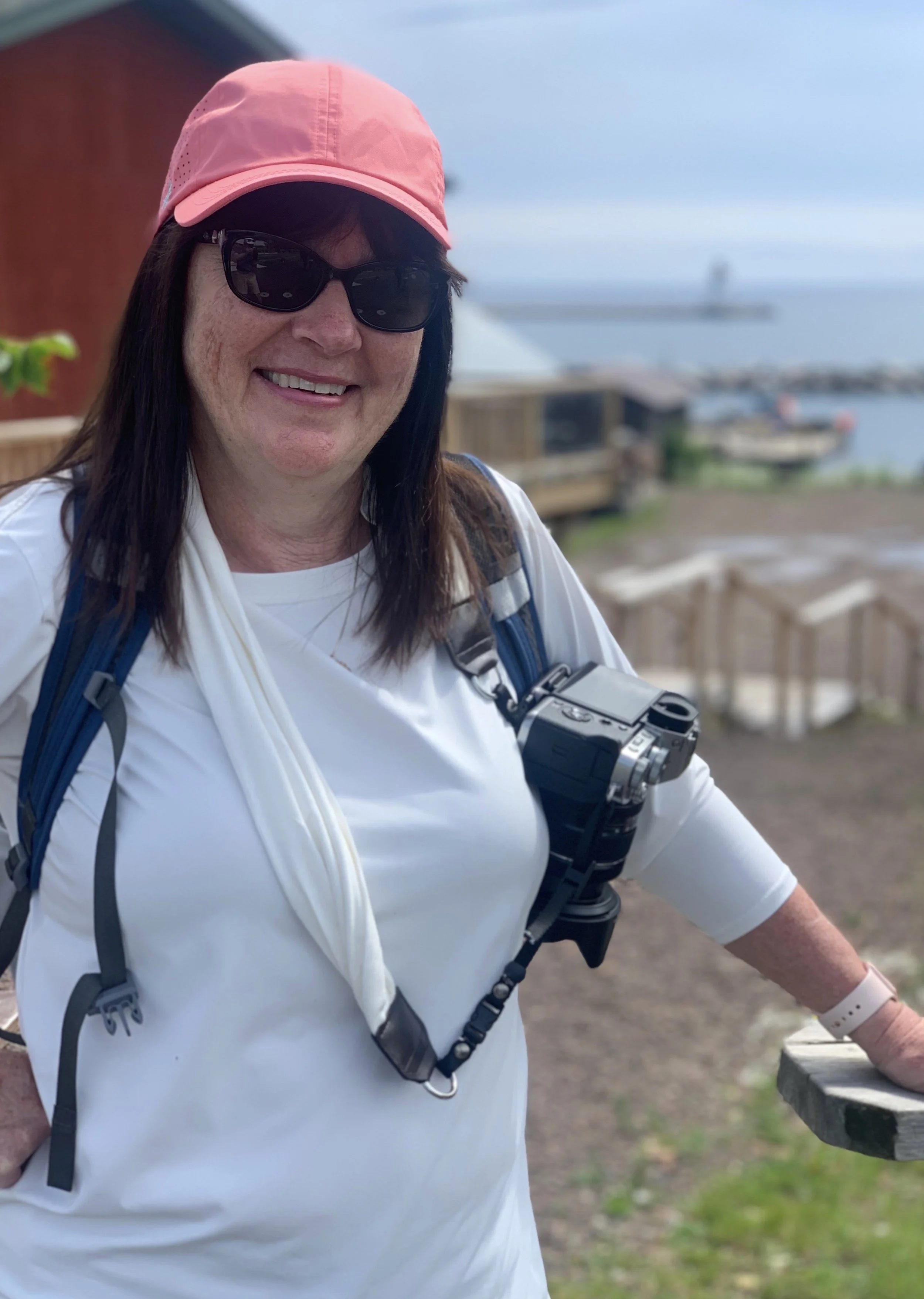 A smiling woman wearing sunglasses, a pink cap, and a white shirt, with a camera around her neck, standing outdoors near a waterfront with a boat and a wooden railing.