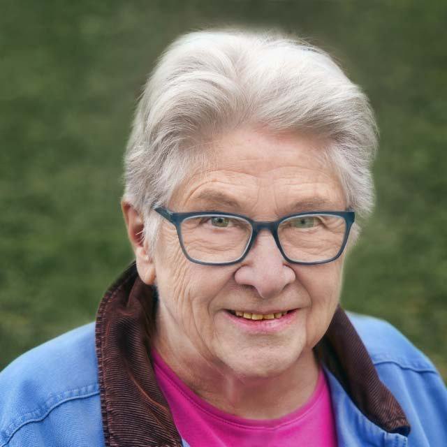 Close-up of an elderly woman with short gray hair, glasses, and a friendly smile, standing outdoors on a grassy background.