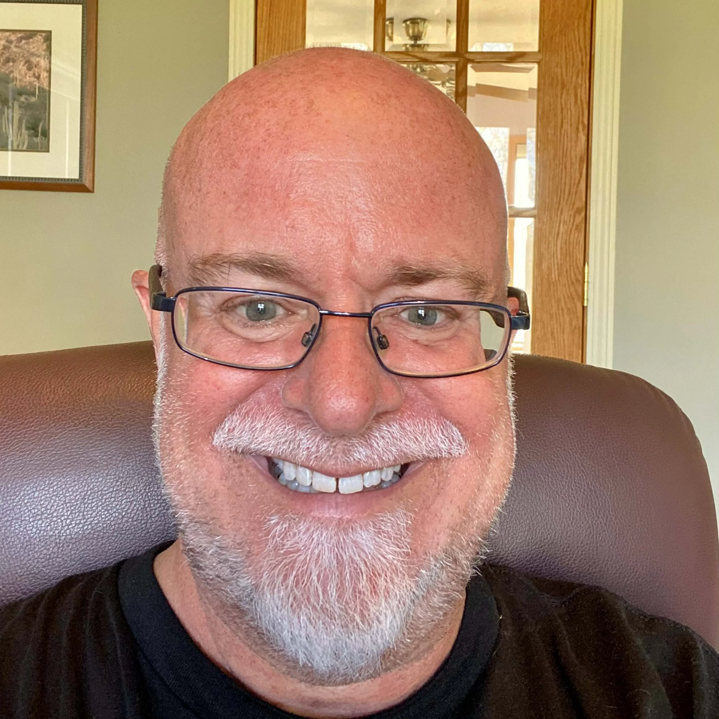 A smiling, bald man with glasses and a white beard sitting on a brown chair indoors, with a framed picture on the wall and a wooden door in the background.