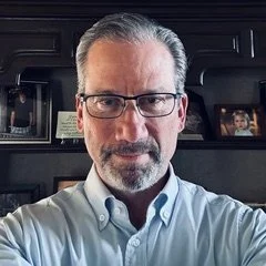 A middle-aged man with gray hair and glasses taking a selfie in front of a dark bookshelf with framed photos and books.