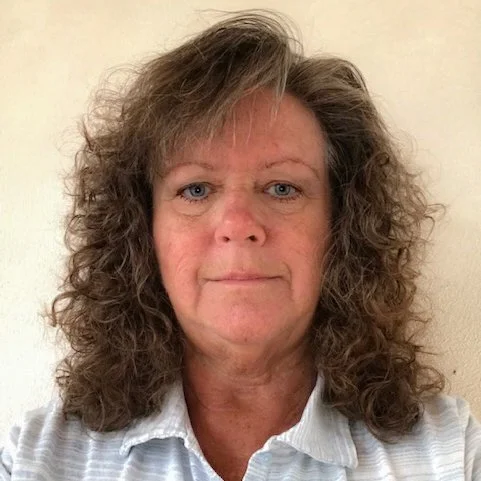 A middle-aged woman with curly, shoulder-length brown hair and wearing a light-colored collared shirt, standing against a plain wall.