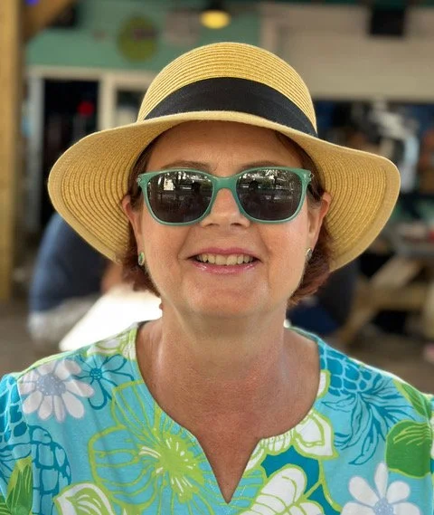 Smiling woman wearing a yellow sunhat, teal sunglasses, and a blue floral shirt in an indoor setting.