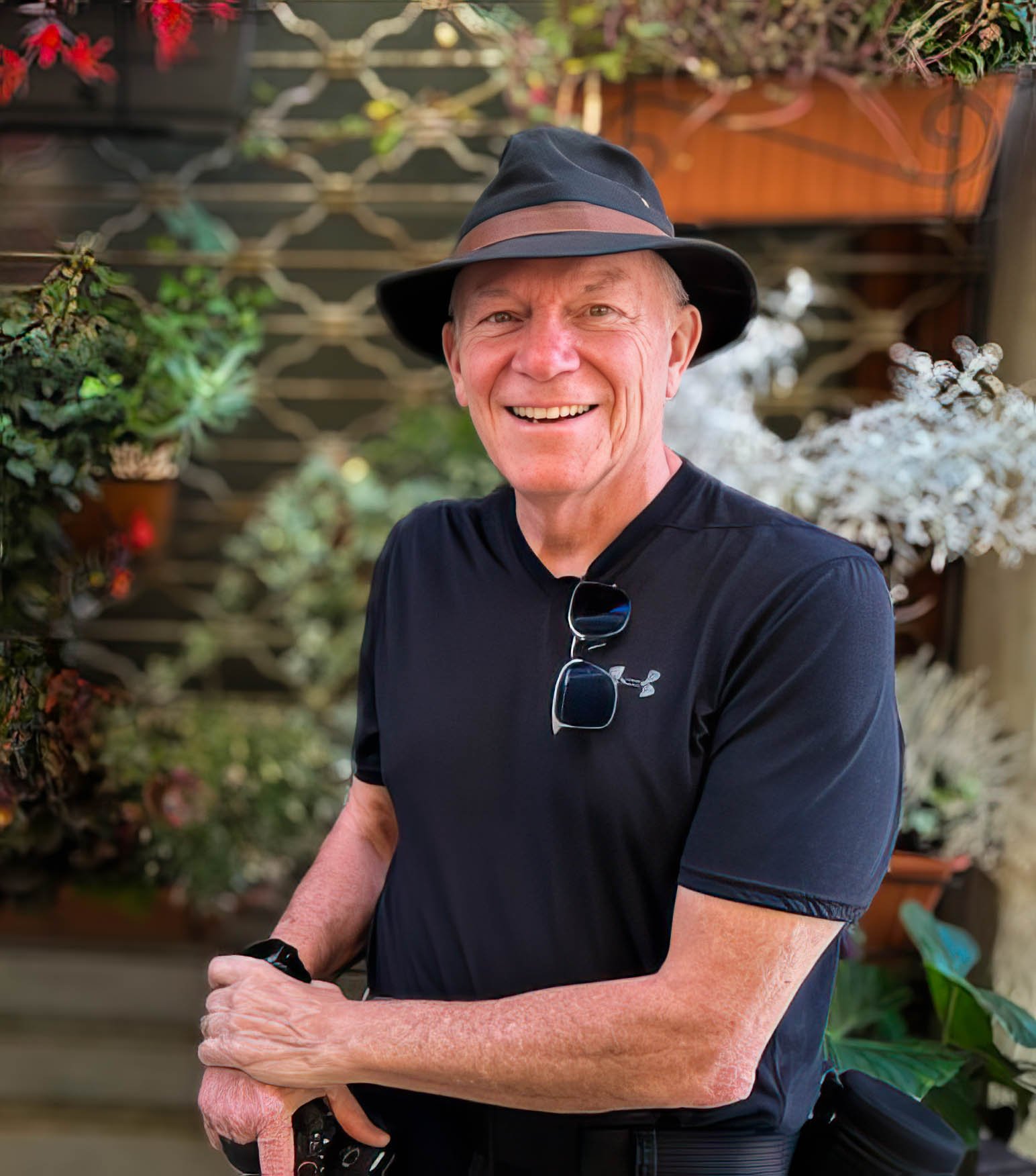 A smiling man with sunglasses hanging from his shirt collar, wearing a black under armor T-shirt and a black hat with a brown band, standing in front of a garden with green plants and flowers.