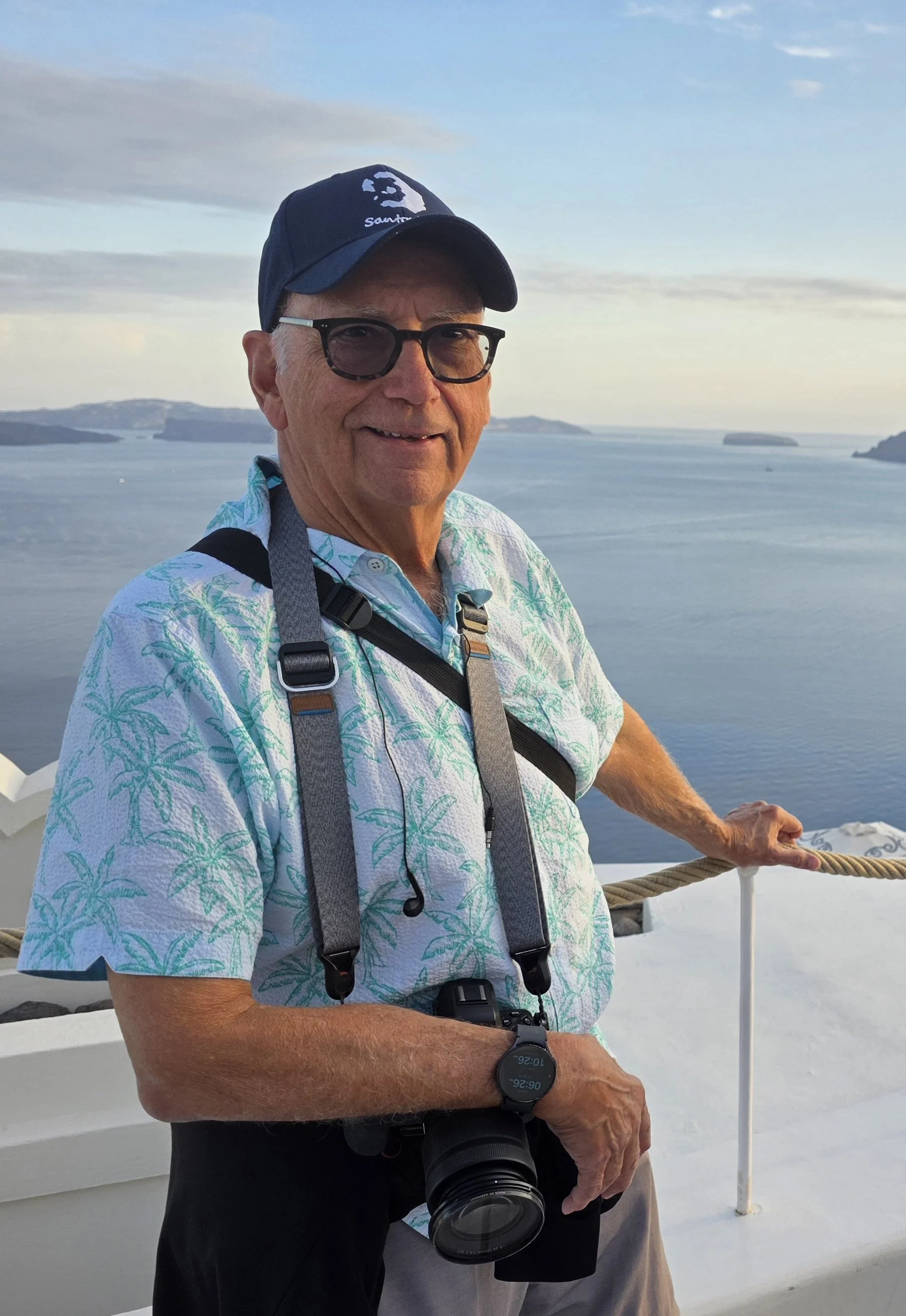 An older man with glasses, wearing a tropical shirt, cap, and camera, standing on a boat or dock with a body of water and distant islands in the background.