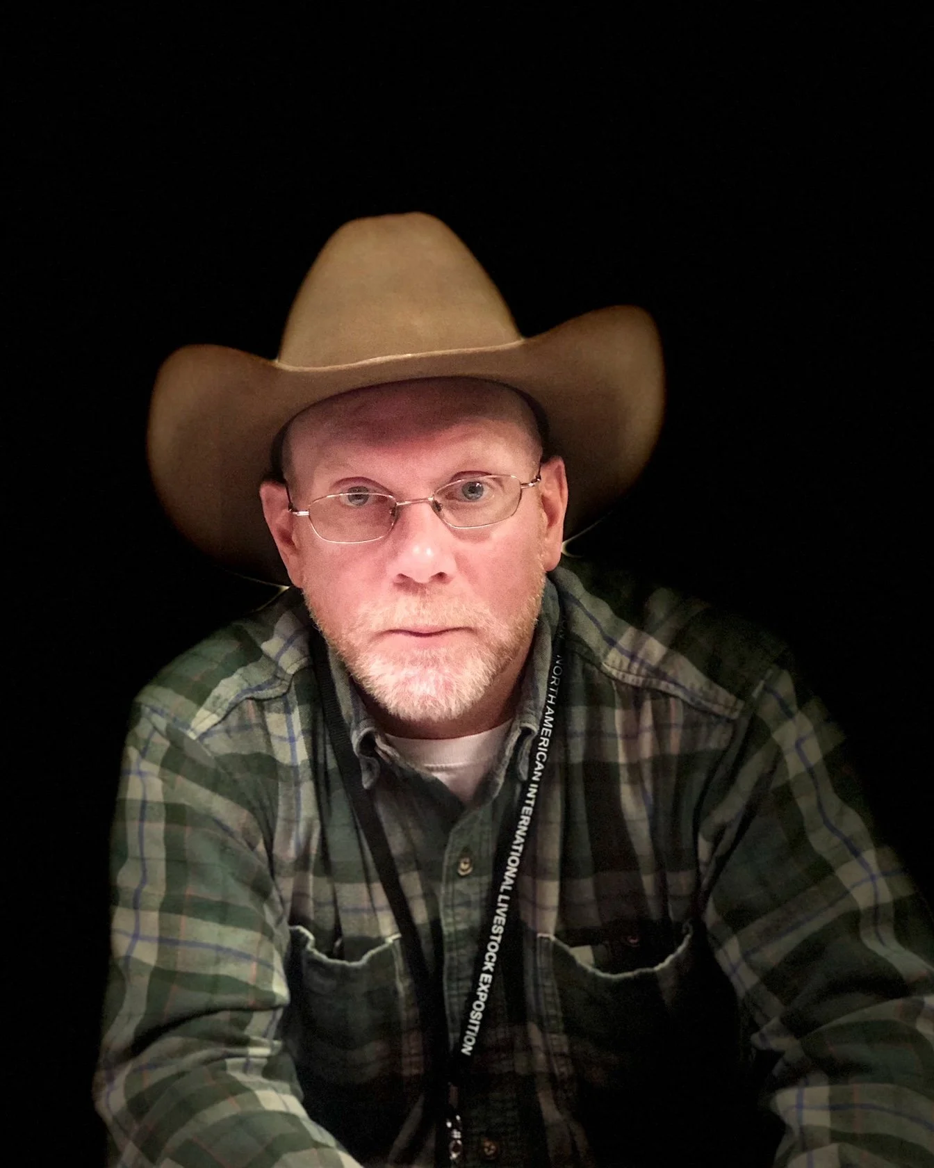 A man with glasses, a beard, and a plaid shirt wearing a large cowboy hat takes a selfie against a black background.