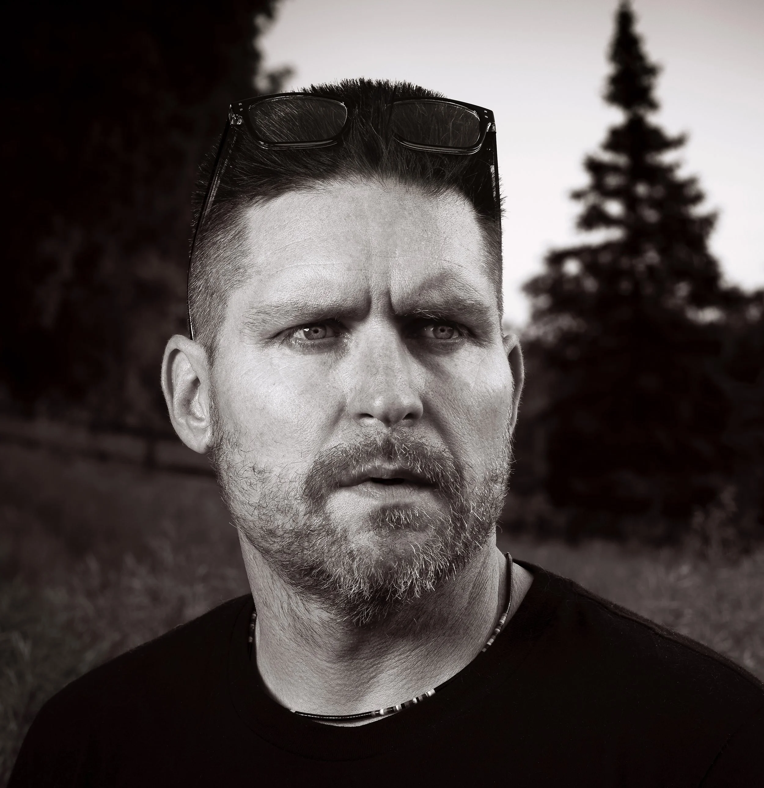 A black and white close-up portrait of a man with short hair, a beard, and glasses resting on his head, looking into the distance with a serious expression, outside with trees in the background.