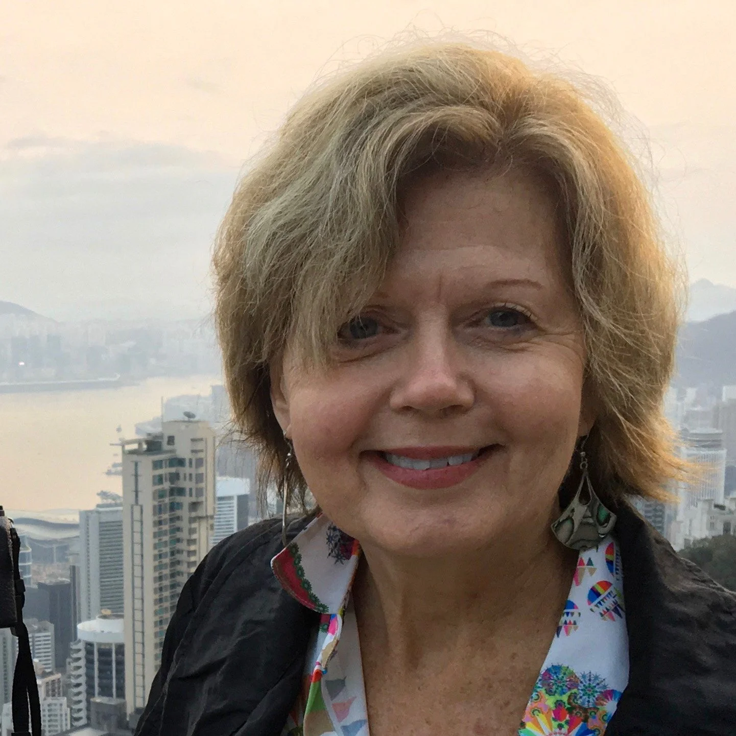 A woman with shoulder-length blond hair smiling outdoors with a city skyline and water in the background.