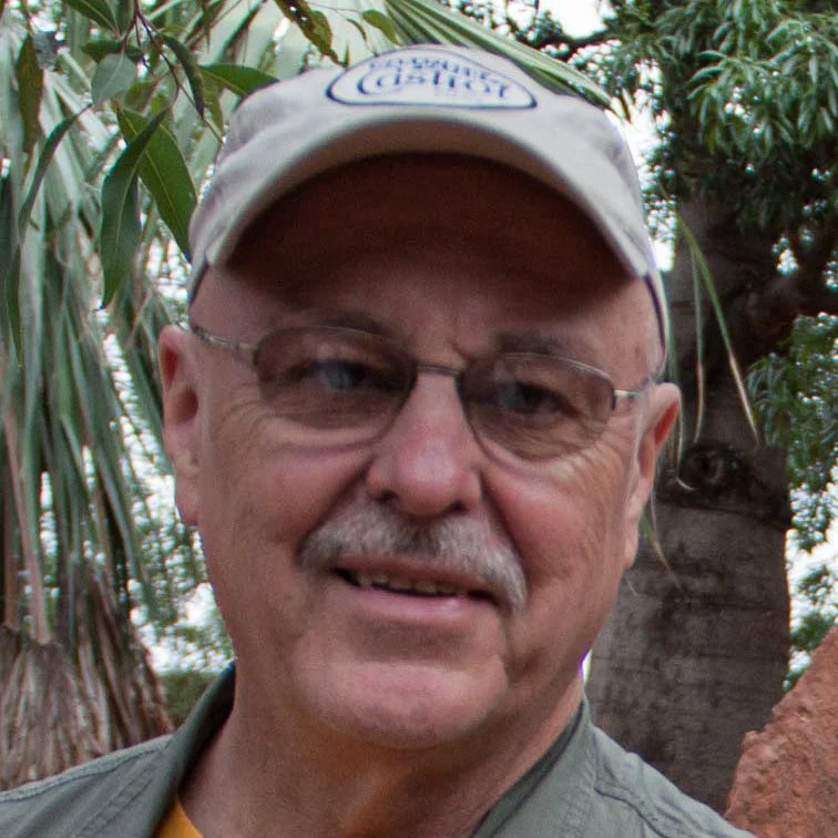 An older man with glasses, a mustache, and a cap, smiling outdoors with trees in the background.