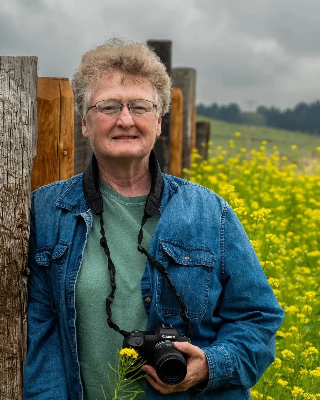 A man with glasses holding a Canon camera stands outdoors in a field of yellow flowers, wearing a denim jacket and green t-shirt, with a cloudy sky in the background.