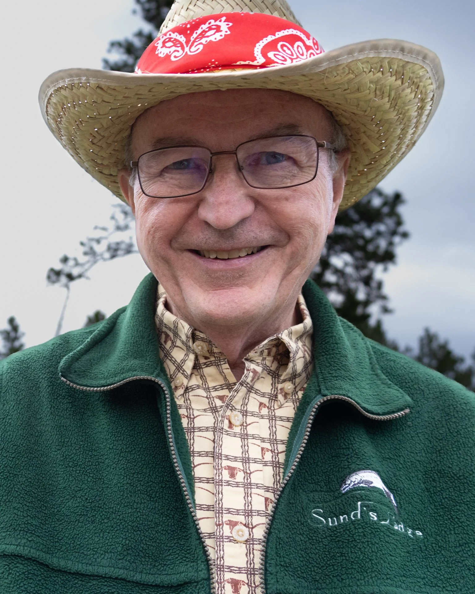 Smiling man wearing a straw cowboy hat with a red bandana, glasses, a green fleece jacket, and a plaid shirt, outdoors with trees in the background.