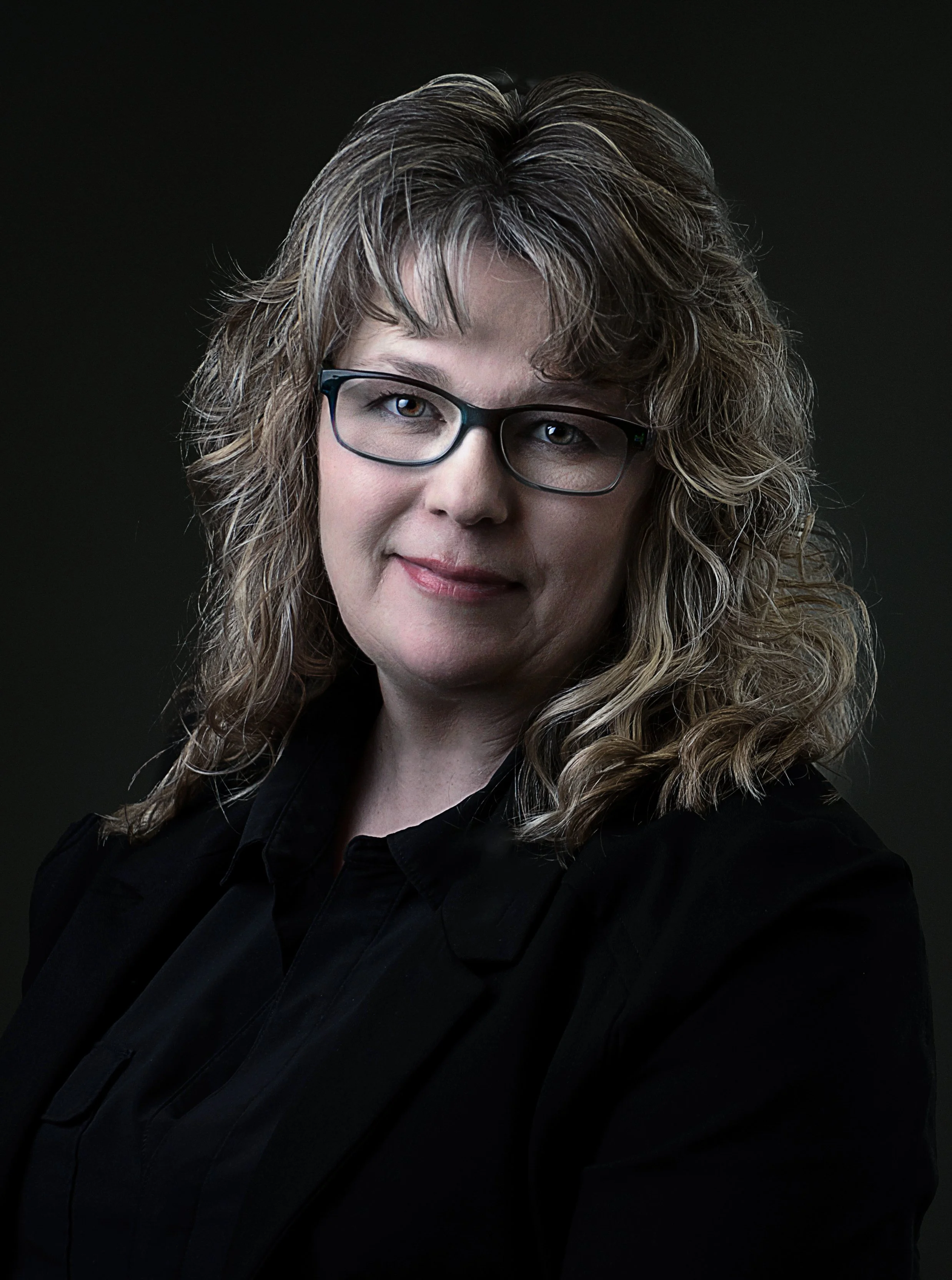 A woman with wavy blonde hair, glasses, and a black shirt, looking at the camera against a dark background.