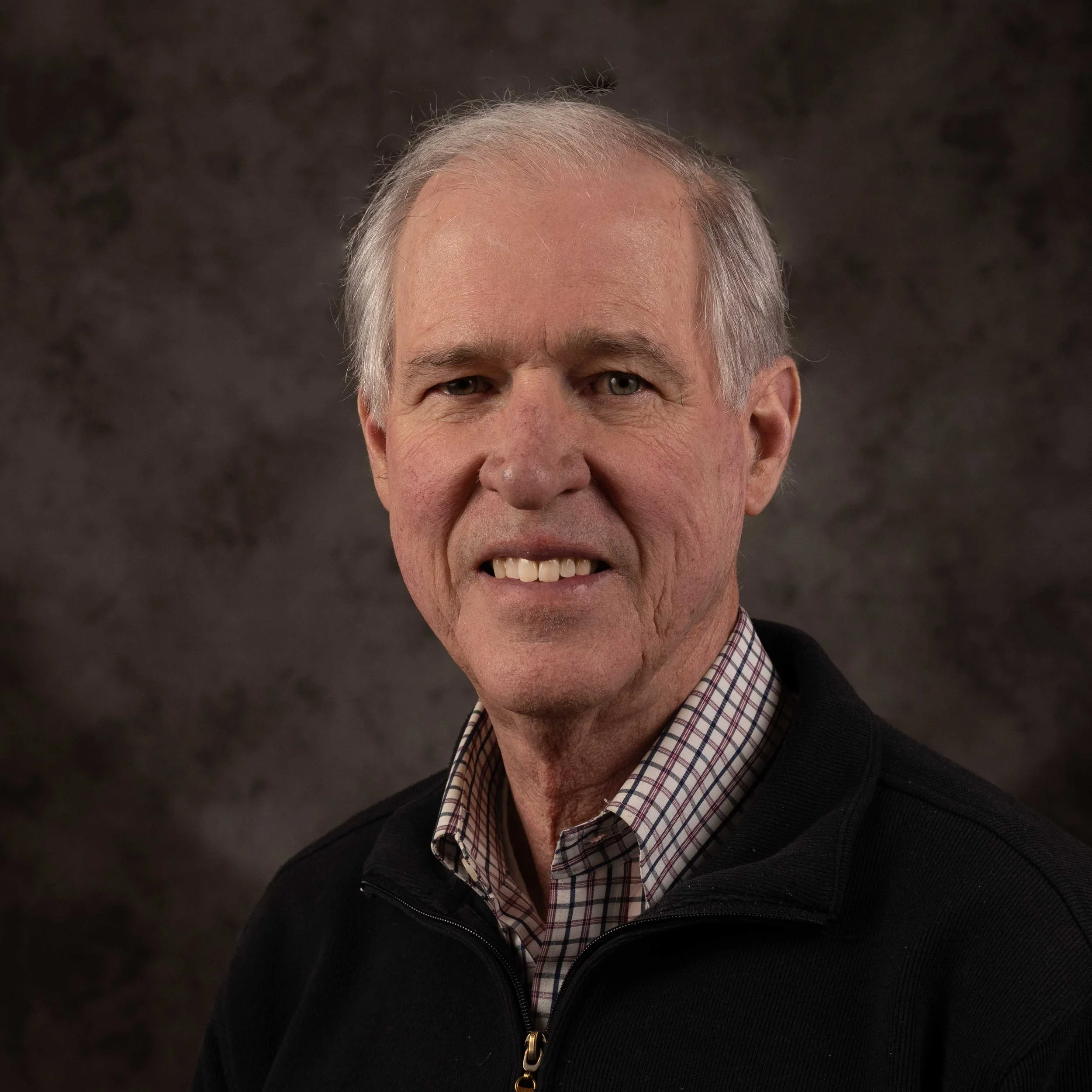 Portrait of an older man with gray hair, wearing a checkered shirt and black jacket, smiling slightly against a dark background.