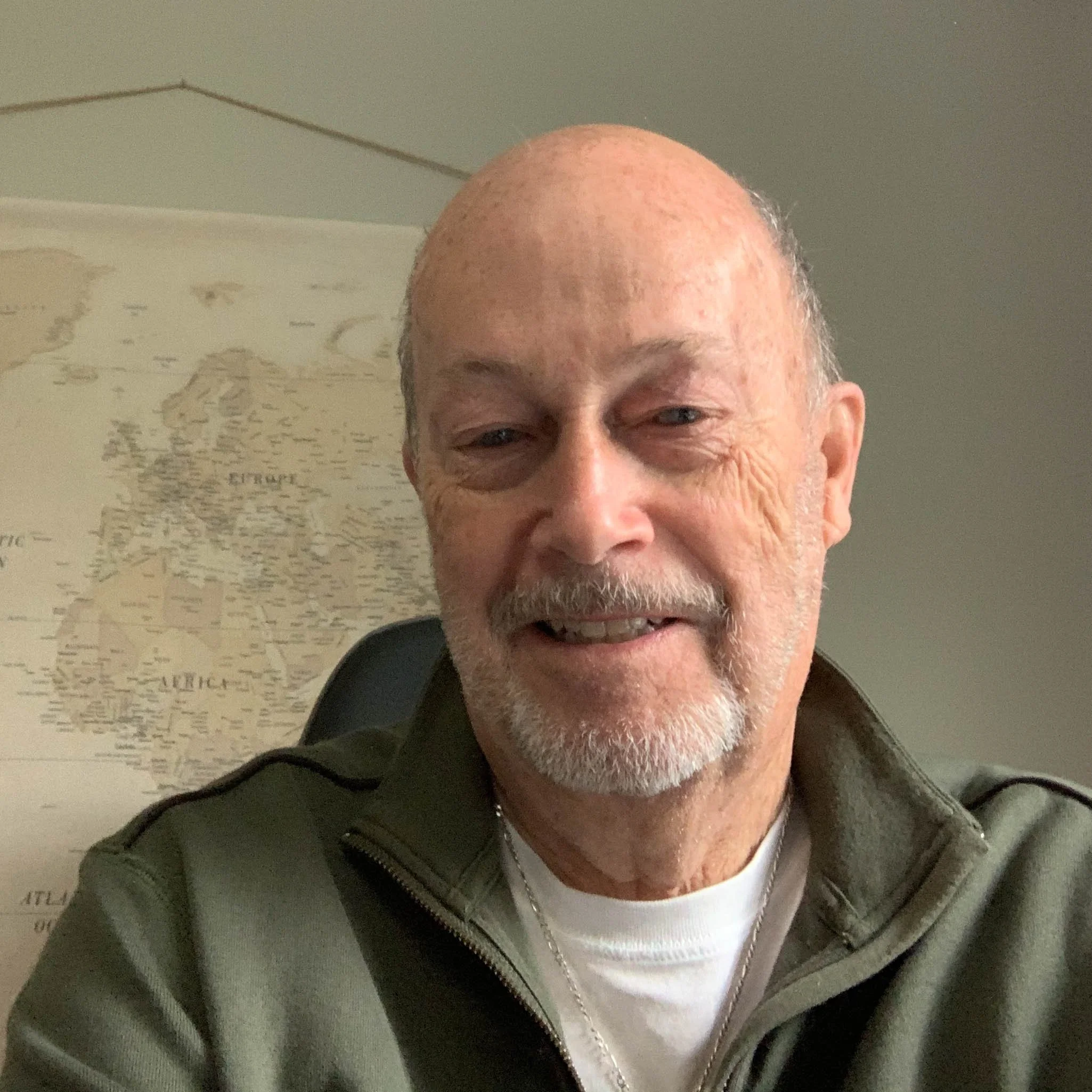 A smiling elderly man with a beard and mustache, wearing a green zip-up jacket and a white shirt, sitting in front of a map of the world on the wall.
