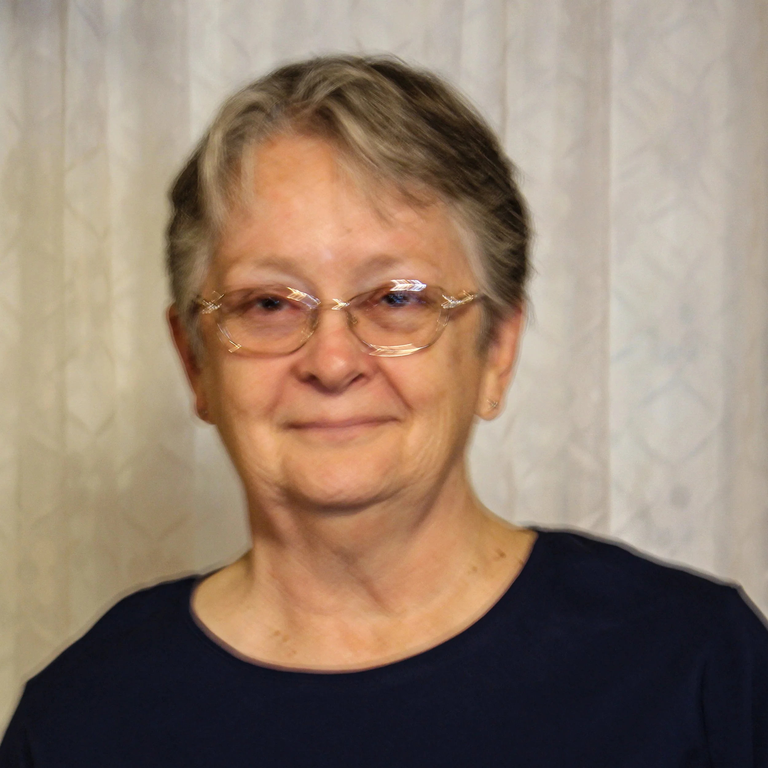 A smiling elderly woman with short gray hair, glasses, and a navy blue top standing in front of a light-colored curtain.