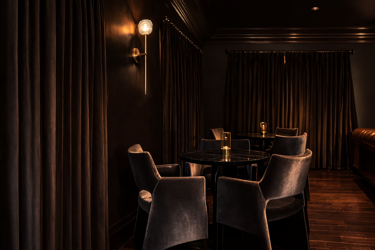 Dimly lit luxurious lounge with dark velvet curtains, a round marble table, and velvet armchairs, illuminated by small table lamps and a wall sconce.