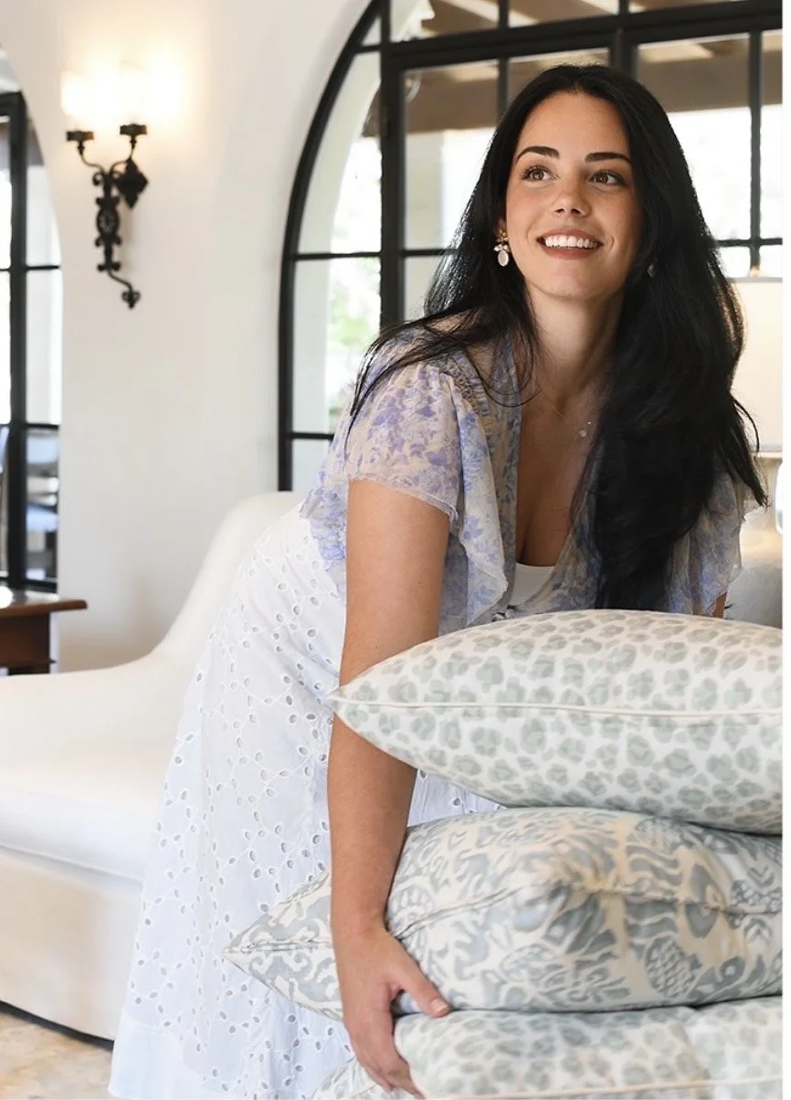 A woman with long black hair, wearing earrings, is smiling and stacking pillows with a floral pattern. She is indoors with large windows and a white sofa in the background.