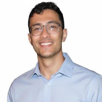 A young man with glasses and short dark hair smiling, wearing a light blue button-up shirt.
