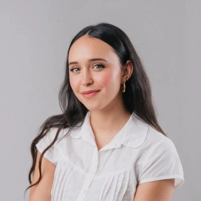 A young woman with long dark hair, wearing a white blouse, smiling slightly, standing against a gray background.