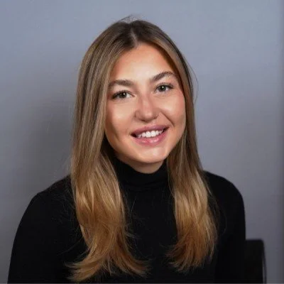 A young woman with long, light brown hair smiling against a plain gray background.