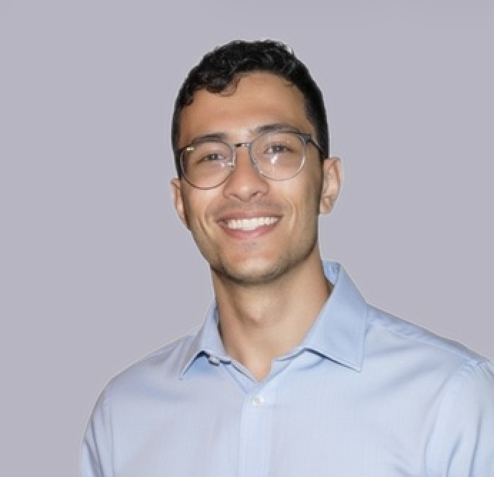 A young man with dark hair, wearing glasses, and a light blue collared shirt, smiling against a gray background.