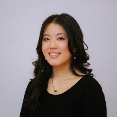 Portrait of a young woman with long black hair wearing a black top and a green pendant necklace, smiling against a plain light background.
