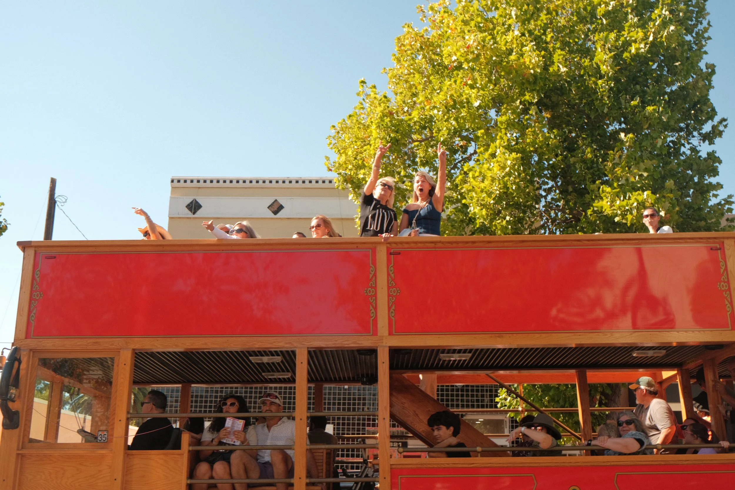 People enjoying a ride on a double-decker bus with open air seating, some taking photos and others pointing or singing, with green trees and a building in the background.
