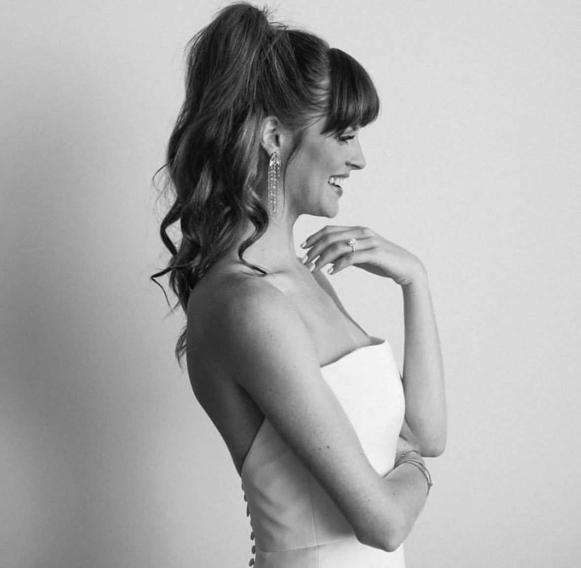 A woman in a strapless dress with wavy hair and earrings, smiling and looking to the side, in a black-and-white photo.