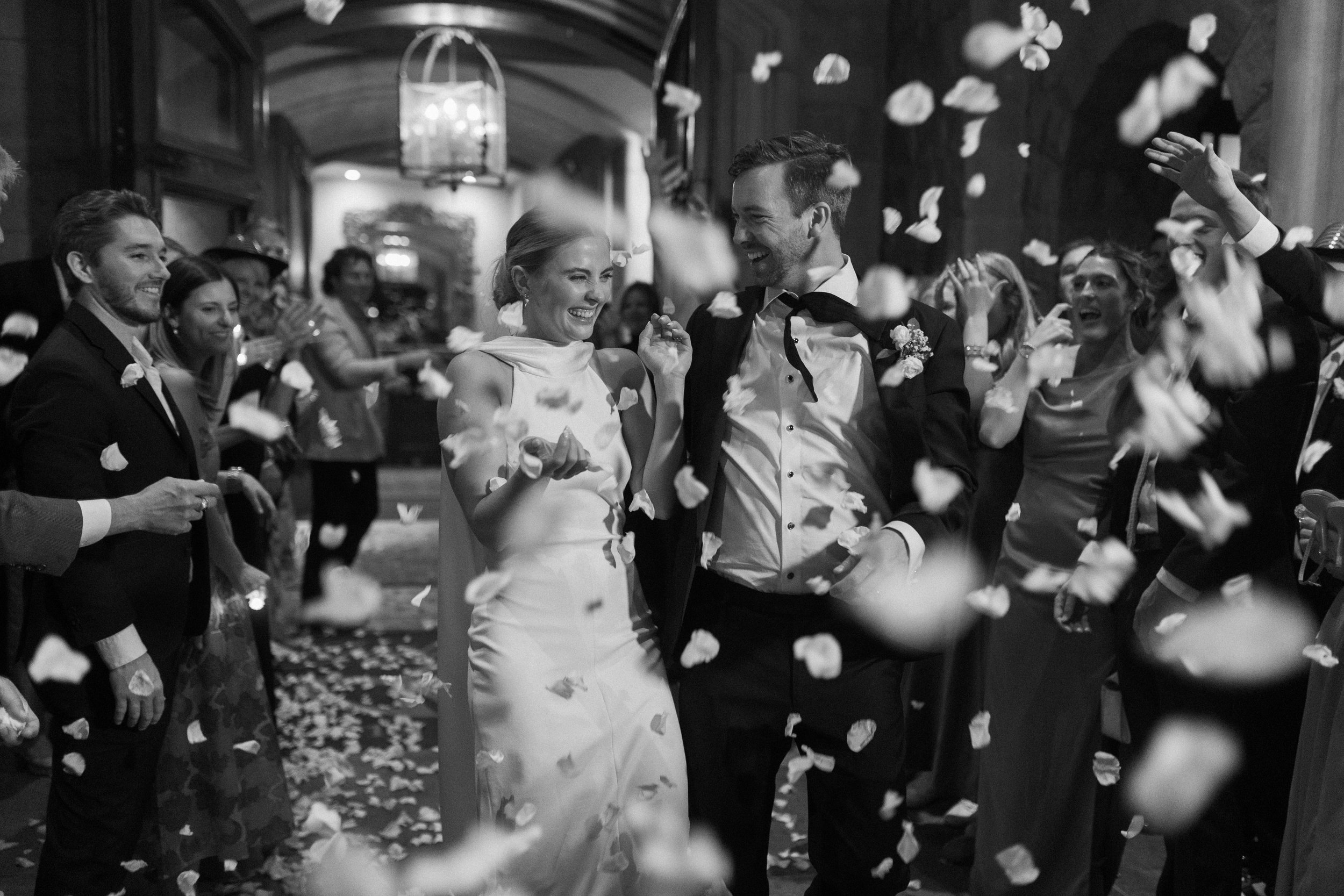 Black and white pic of a wedding celebration with a woman and a man dancing among guests, with white flower petals falling in the air, in an elegant venue.