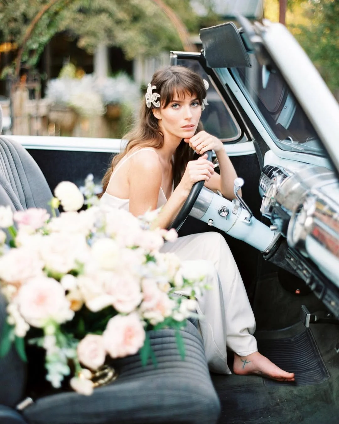 A woman with long brown hair, wearing a white dress and floral hair accessories, sitting in an open vintage car with a bouquet of pink and white flowers on the front seat, outdoors with trees in the background.