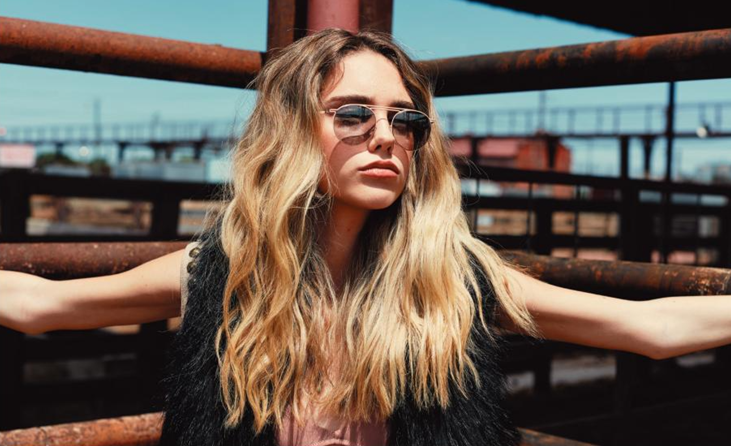 A young woman with long, wavy blonde hair wearing sunglasses and a black vest, standing outdoors on a metal structure with a cityscape in the background.