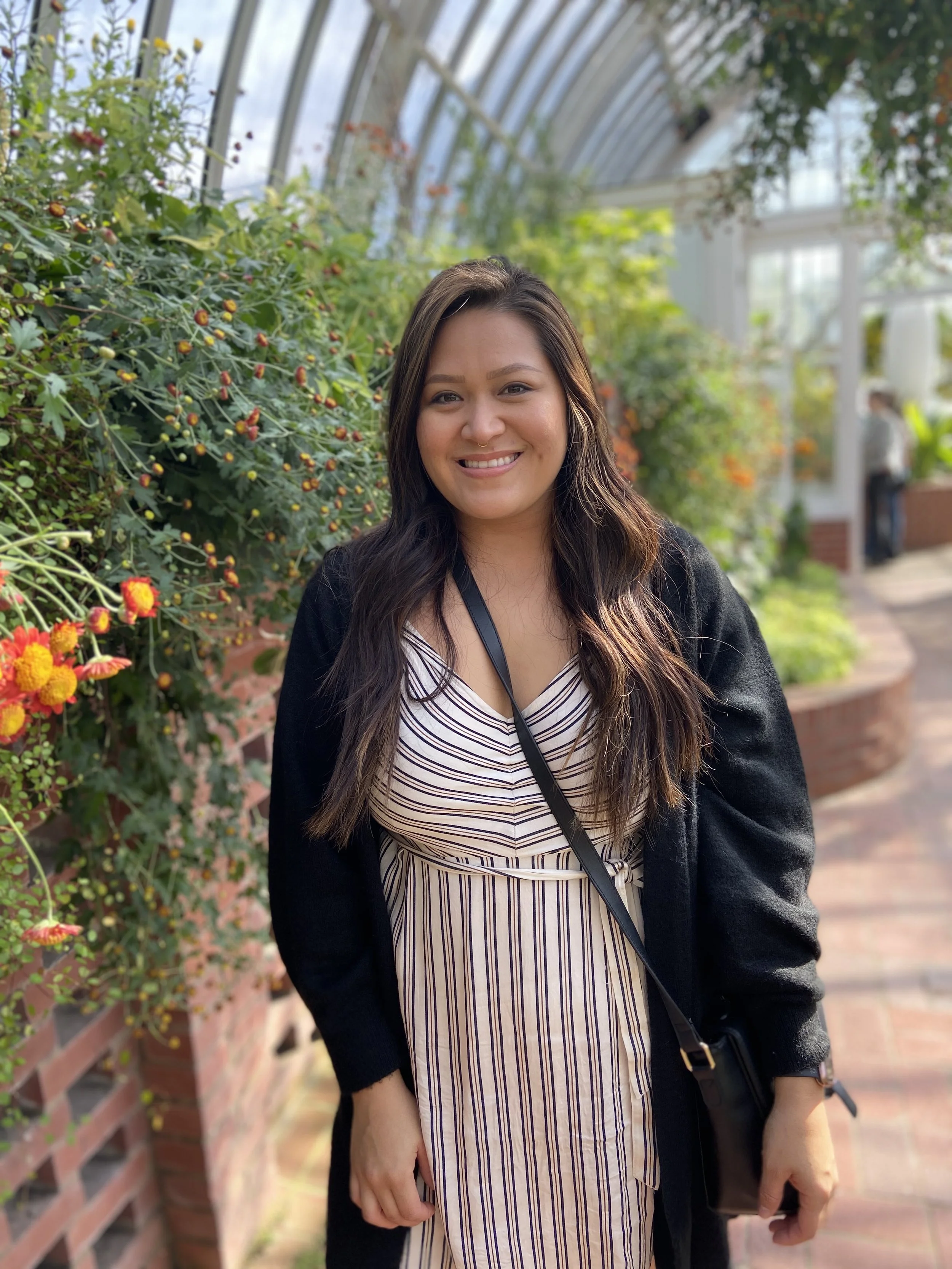A woman with long dark hair smiling and standing in a greenhouse or conservatory filled with lush green plants and colorful flowers.