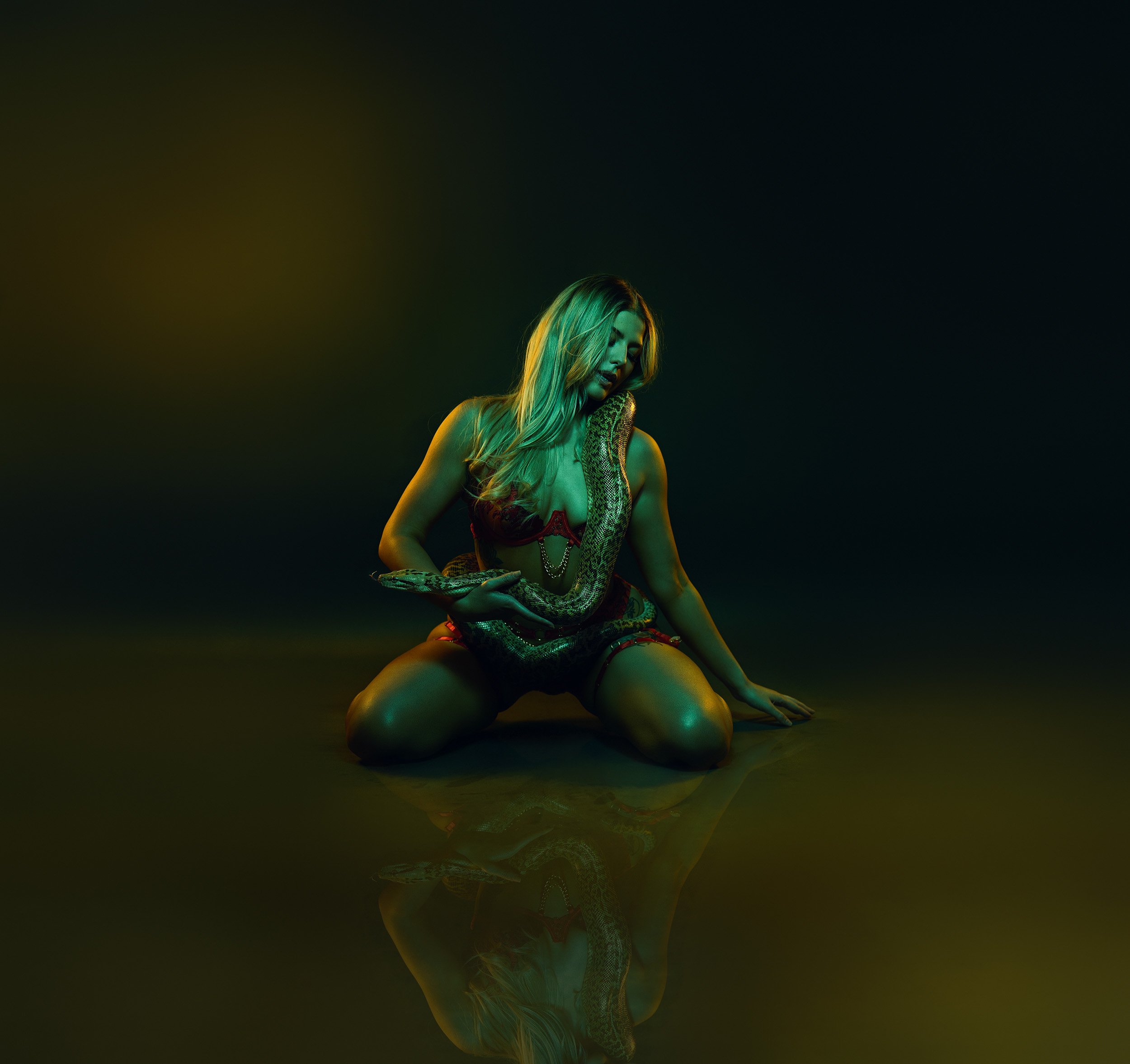 Luxury boudoir portrait of a woman kneeling in black lingerie on a reflective floor with moody green and gold lighting.