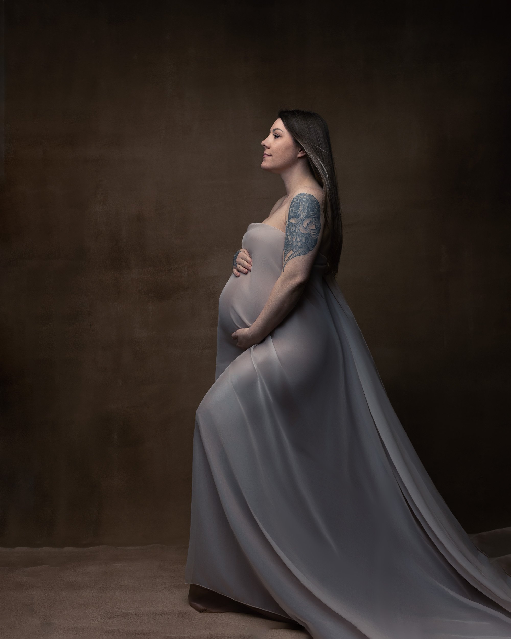 Fine art maternity portrait by Amanda Chantee in Vancouver, WA, featuring a tattooed pregnant woman in a flowing silver gown against a warm painterly backdrop.